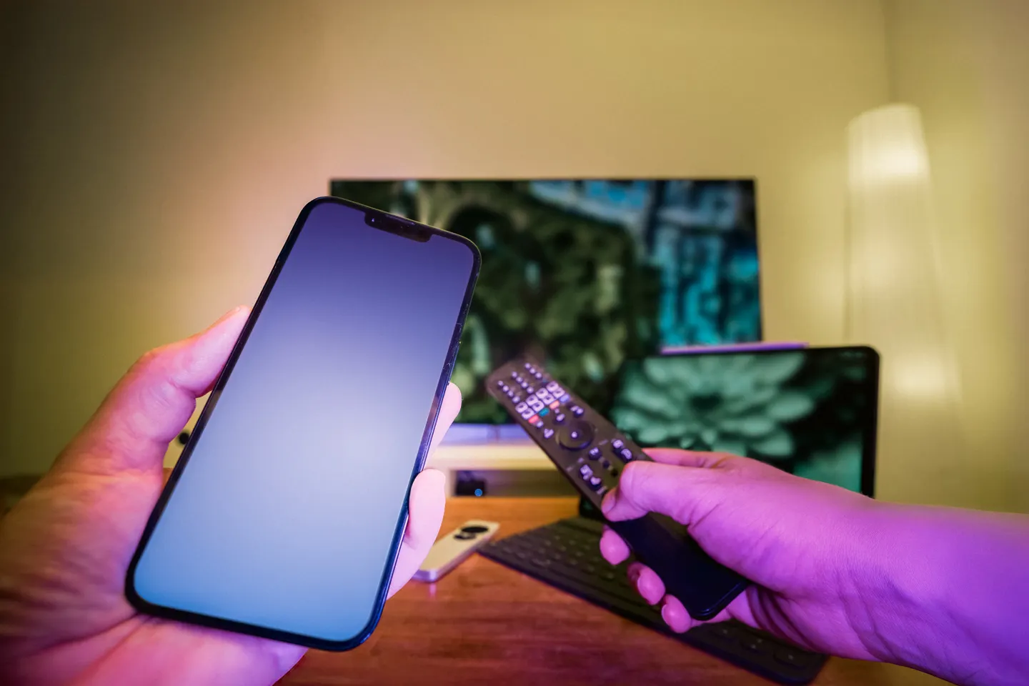 Handen met een smartphone en afstandsbediening voor een televisie en laptop